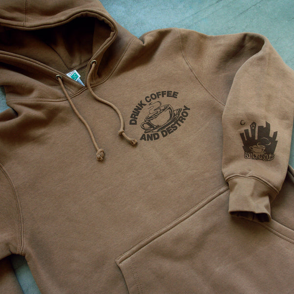 close up angled image of a toasted coconut colored pullover hoodie laid flat on a concrete floor. small brown right chest print of a cup of hot coffee. above it says drink coffee, and below it says and destroy. there is a small cuff print on the bottom right sleeve of a city scape and says bouncing souls