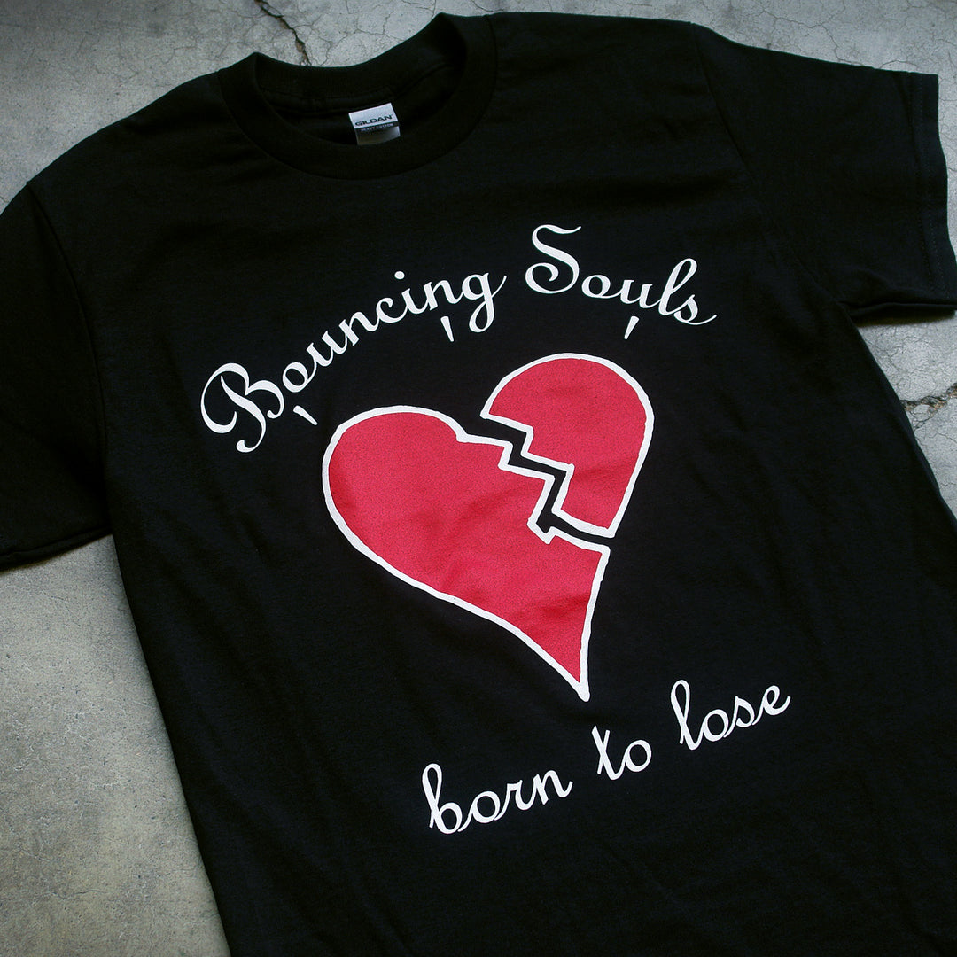 image of a black tee shirt on a concrete background. the front of the tee has a full chest print.the top of the shirt says bouncing souls with a broken red heart in the middle and born to lose below the heart.